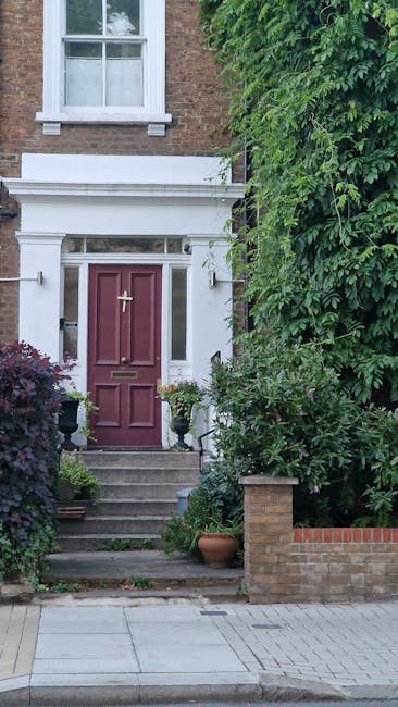 A narrow garden pathway between a white exterior wall of a house and dense, leafy shrubbery on the left side, with textured wet soil and weathered paving stones. The pathway is flanked by a few potted